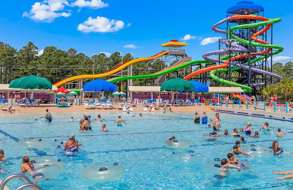 a large outdoor pool with people swimming and playing in the water, with a water slide in the background